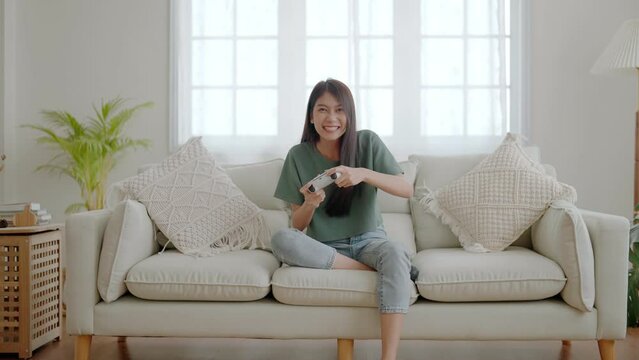 Beautiful young asian woman sitting on sofa and holding joystick playing online video game in living room at house