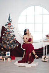 Portrait of pretty young girl cozy sit down and drinking wine, wear red gown in decorated Christmas living room indoors