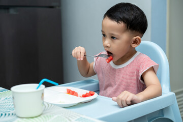 Happy toddler boy eating watermelon his high chair in dining room