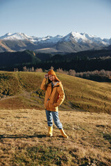 Fototapeta na wymiar Autumn woman walking up the hill in full smile travels to the mountains in nature hiking and happiness in a yellow cape against the snowy mountains in the sunset, freedom of life style
