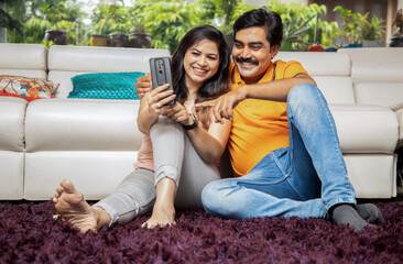 Portrait of happy young indian couple using smart phone while sitting on floor together at home. Asian Man and Woman holding Mobile Phone taking selfie picture, Full Length Shot.
