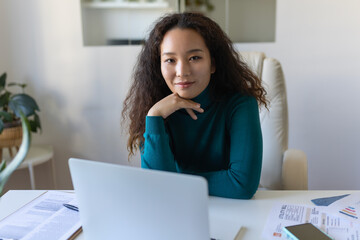 Shot of Asian smiling business woman working with laptop while looking at camera in modern startup office.