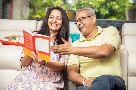 Happy senior indian couple reading book or novel together at home. 60s Retired husband wife spend time with each other and having fun. Retirement life.