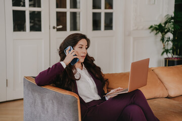 Focused brunette businesswoman in violet suit sitting on leather couch using laptop talking by phone at home. Remote working hispanic lawyer consulting client via internet. Business and finance.