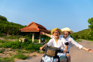 Happy Asian family couple enjoy and fun outdoor lifestyle on summer beach holiday vacation. Husband and wife riding motorcycle together while travel mountain road on tropical island in sunny day.