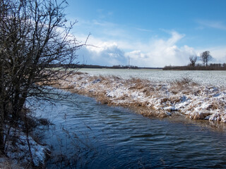 Landscape of a big ditch full with water surrounded with big fields covered with snow in winter