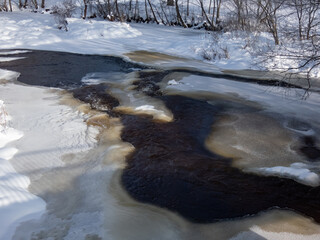 View of a big river partly frozen with visible flowing, dark water and banks and surroundings covered with snow in winter