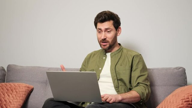 Handsome Berded Busy Man Working on Laptop Computer Having Video Call Distantly while Sitting on a Sofa in Cozy Living Room. Freelancer Working From Home. Browsing Internet, Using Social Networks.