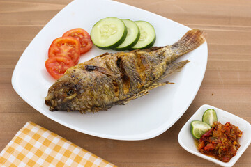 Close-up of Fried Nile Tilapia fish, Indonesian traditional food. Served on white plate with sambal (chilli sauce), cucumber and tomato slic