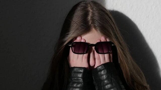 Girl in a leather jacket and sunglasses, covers her face with her hands, an upset child