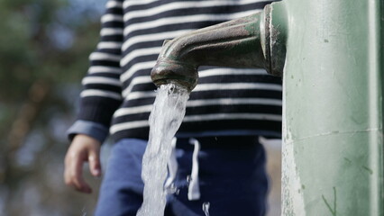 Closeup of public faucet flowing water at park outdoors with child standing in background