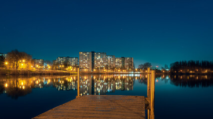 Fototapeta premium Night view on Tysiaclecie estate in Katowice, Silesia, Poland. Lightened residential buildings with surrounding trees at dusk seen through the lake. Little, wooden jetty in the foreground.
