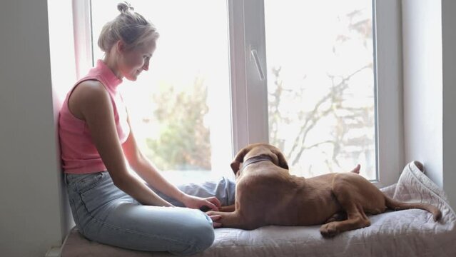 A joyful girl sits with a puppy of Bordeaux dog on the windowsill. The concept of love and care for pets