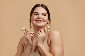 Face massage. Plus Size Woman Holding Jade Facial Roller for Skin Care, Beauty Treatment on Beige Isolate Background. Girl Taking Care of Skin with Natural Massager Closeup Portrait 