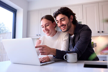 Fototapeta na wymiar Couple At Home Looking At Laptop On Counter In Kitchen Together