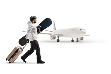 Young man holding a snowboard and pulling a suitcase in front of an airplane