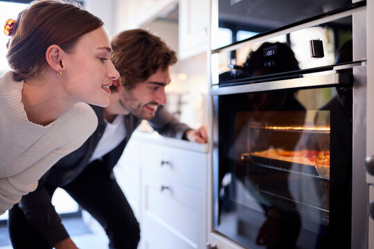 Couple In Kitchen At Home Watching Homemade Pizza In Oven Baking