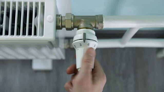 Man regulates the heat supply to a radiator with a thermostat knob. Hot pipes in winter. Increasing and decreasing the temperature in the system.