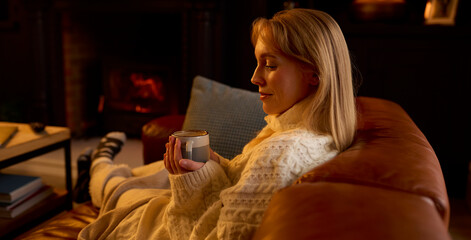Woman At Home In Lounge With Cosy Fire With Hot Drink