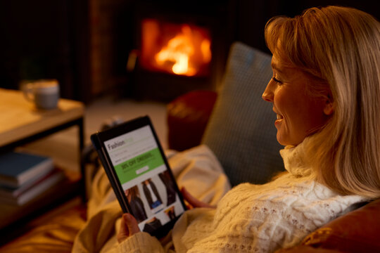 Woman At Home In Lounge With Cosy Fire Looking At Fashion Website On Digital Tablet