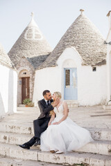 Happy blissful wedding couple sitting on stairs near conical roofs of trulli, kissing. Young bride...