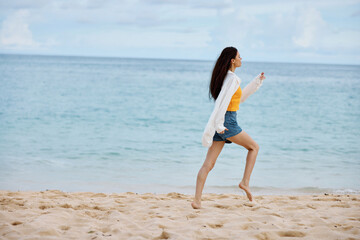 Sports woman runs along the beach in summer clothes on the sand in a yellow tank top and denim shorts white shirt flying hair ocean view, beach vacation and travel