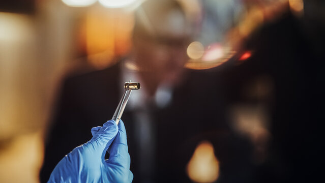 Extreme Close Up with Blurry Background: Hand with Medical Glove Using Tweezers to Hold a Bullet Shell. Forensics Specialist Finding Cartridge on Crime Scene and Showing it to Detective as Evidence