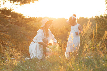 Mother with little daughter sit in white dresses on high hill in wood among thick grass at yellow...