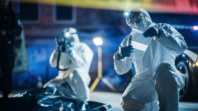 Portrait of Two Forensics Experts Doing Fieldwork at Night at a Crime Scene. One Technician Taking Photos of the Dead Body While the Other Packs the Bloodied Knife as Murder Weapon.