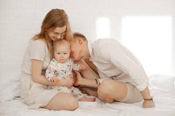 Fototapeta na wymiar Portrait of happy family of three on white background. Mother embrace and father with love kiss infant baby. Photo of young couple and child in white family look. Parental affection, childhood