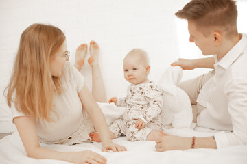 Obraz na płótnie Canvas Portrait of happy family of three on white background. Mother, father and infant baby lie on bed. Photo of young couple and child in white family look. Parental affection, childhood, love