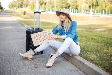 Solitude, upset melancholy blond woman in hat hitchhiking, sit on curb by road, hold carton board anywhere with suitcase