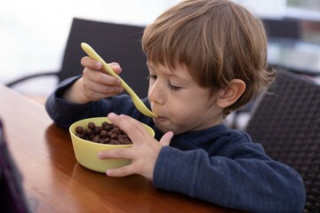 Hungry little boy sitting at wooden table in kitchen and having breakfast closeup. Kid of kindergarten age holding spoon to eat dry chocolate balls without milk. Ready breakfast, health nutrition