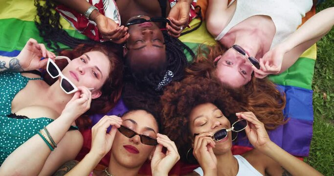 High Angle: Group of Young Multiethnic Women Putting on Sunglasses and Posing for a Selfie On Smartphone While Lying Down on a Rainbow Flag. Female Diverse Friends Having Fun in a Park. Lgbtq+ Allies
