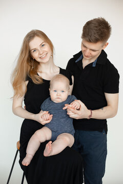 Happy family from three in black looks on white background. Mom with beautiful smile sit on chair and hold infant baby, dad stand near. Family photo, parenting, firstborn, love, feelings