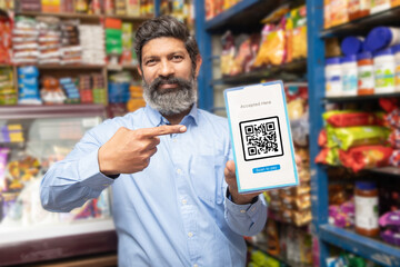 Portrait of happy mature Indian man showing Qr Code scanner to pay online at grocery shop or supermarket. Cashless payment. - obrazy, fototapety, plakaty