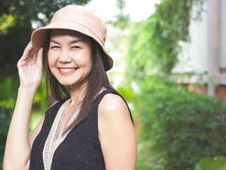 Asian woman, wearing hat and black top sleeveless, standing in the garden, touching her hat,  smiling happily and looking at camera.