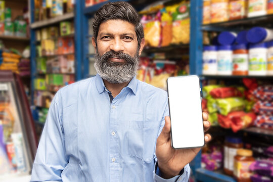 Happy Indian man small business owner show smart phone with blank display screen to put advertisement at grocery shop or supermarket,