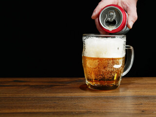 Pouring beer into big glass jar on a brown wooden table. Alcohol consumption. Dark background.