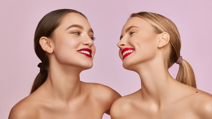 Red lipstick beauty. Portrait of two young, beautiful girls with well-kept skin isolated over pink studio background. Concept of skincare, cosmetology, natural beauty