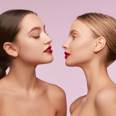 Red lips. Portrait of two young, beautiful girls with well-kept skin isolated over pink studio background. Concept of skincare, cosmetology, natural beauty