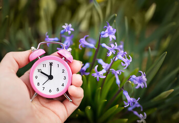 Hand holding an alarm clock on purple blooming easter flowers. Spring forward, springtime. Daylight savings time.