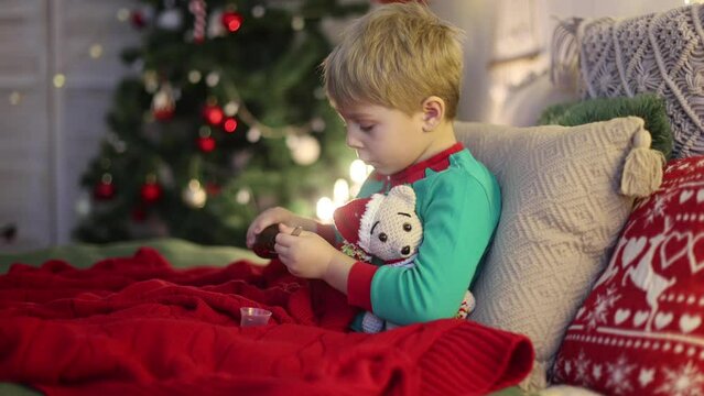 Toddler child, cute blond boy, sitting on the window in pajama, looking out for Satna Claus,, eating cookies,  Christmas lights around him