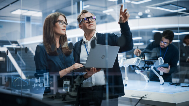 Portrait Of Two Young Female And Male Engineers Use Laptop Computer To Connect, Analyze And Discuss How To Program A Robot Dog. Standing In High Tech Research Laboratory With Modern Equipment.