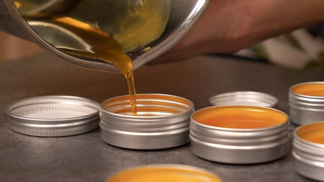 Close-up shot of pouring a hot yellow-orange liquid beeswax balm into an empty circular aluminum container. DIY Homemade Organic Lip Balm.