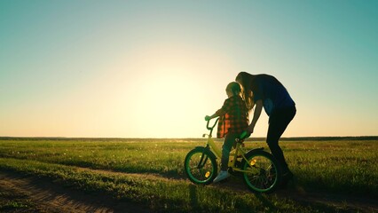 Active Mom teaches her little daughter to ride bike, sunset. Mother teaches her child to keep balance while sitting on bicycle. Childhood dream of riding bike. Family life, mom, baby, parental support
