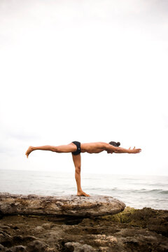 A fit man performs a difficult balancing pose while practicing yoga on a rocky beach.