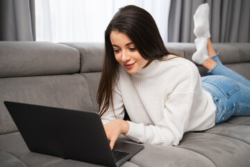 Fototapeta premium Young female laying on the sofa and using her notebook. A smiling woman has online shopping using a laptop at home. Working from home as a freelancer.