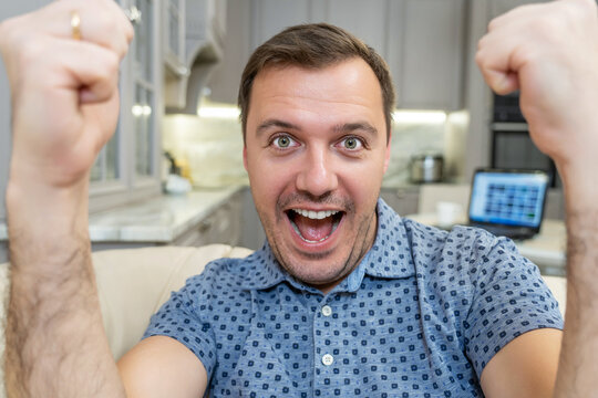 Millenial Man At Home Alone Watching Games On Tv Enjoying And Celebrates Goal And Gestures Of Victory At Living Room. Happy Man Having Video Call With Friends, Showing New Apartment After Relocation