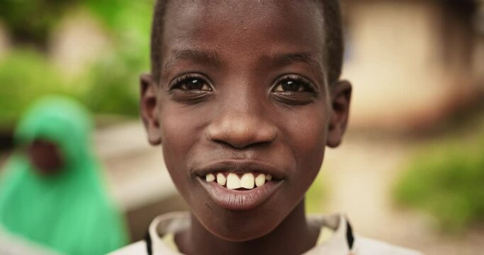 Close Up Portrait of a Shy Authentic African Boy Posing and Smiling at the Camera with a Blurred Rural Village in the Background. Black Male Kid Hopeful about the Future and Solidarity of the World